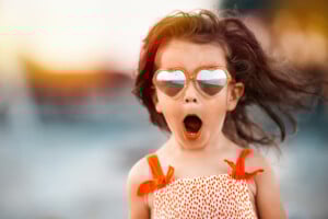 A young girl named Isla, with long hair, wears heart-shaped sunglasses and an orange and white polka dot dress. She appears to be surprised or excited, her mouth open wide. The background is blurred, suggesting an outdoor setting near water.