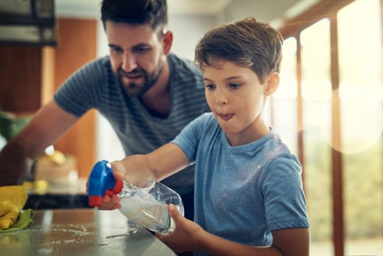 A man and a boy are cleaning a countertop in a kitchen, demonstrating how parents should treat their child as capable little adults. The man watches while the boy sprays cleaner from a bottle onto the surface. Both are dressed casually, and the kitchen is well-lit with natural light.