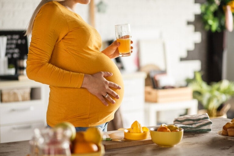 A pregnant woman in a yellow long-sleeve shirt is standing in a kitchen holding a glass of orange juice. On the table in front of her, alongside various fruits and kitchen towels, there's a bottle of castor oil for inducing labor. The background reveals a shelf with plants and kitchen items.