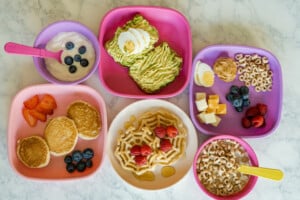 A variety of breakfast foods are served on colorful plates. Items include waffles with strawberries, avocado toast with boiled egg, yogurt with blueberries, cheese cubes with blueberries, pancakes with strawberries and blueberries, and cereal with milk—perfect healthy breakfast ideas for kids.