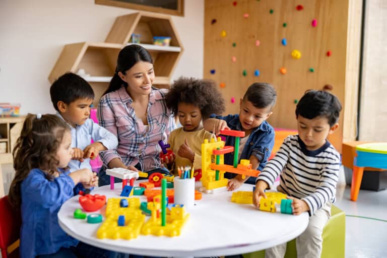 A teacher sits with five children at a round table, engaged in play with colorful building toys in what could be the best daycare setting. Shelves and a wall with climbing holds complement the lively classroom atmosphere, showcasing how to find a good daycare that fosters creativity and joy.