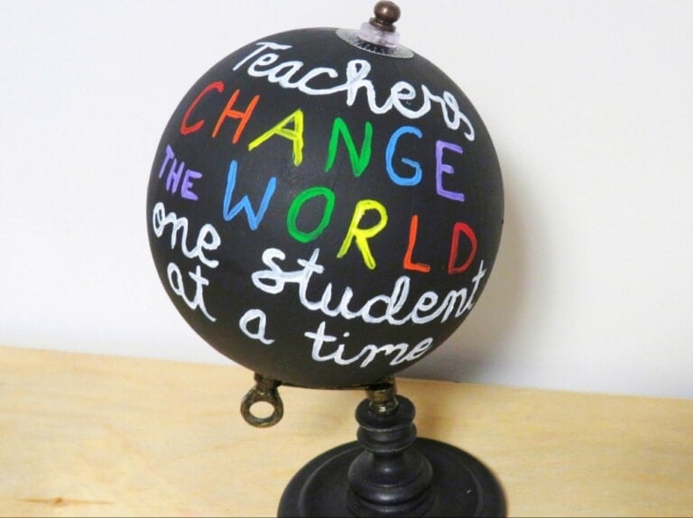 A small black globe on a stand features colorful handwritten words: "Teachers change the world one student at a time." Perfect as one of those thoughtful DIY teacher appreciation gifts, the globe is placed on a light wooden surface against a plain white background.