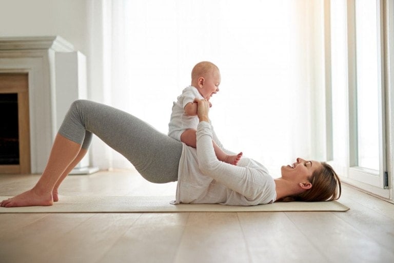 A woman is lying on her back on a yoga mat, lifting her hips in a bridge pose while holding a baby who is sitting on her stomach. Both are smiling, making the most of exercising after a c-section in a brightly lit room with wooden flooring and large windows.