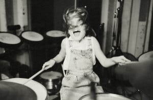 A child with shoulder-length hair is enthusiastically playing the drums, embodying the spirit of badass girl names. Their hair is in motion, covering part of their face as they rock out in overalls and a sleeveless shirt. The background features more drum equipment in this striking black-and-white image.