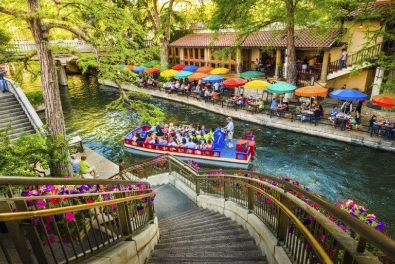 A scenic view of a river with a boat full of passengers floats by a riverside restaurant in San Antonio. Colorful umbrellas shade diners seated at outdoor tables, perfect for a babymoon outing. Flowers adorn the stairway leading down to the water, and trees and greenery create a relaxing atmosphere.
