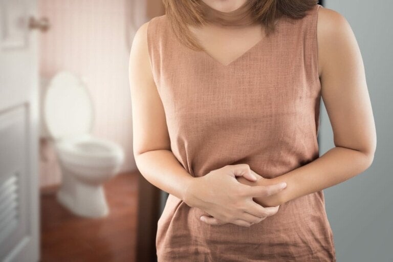 A person in a brown sleeveless top holds their stomach with both hands, indicating discomfort. In the background, a white toilet is visible in a brightly lit bathroom. The setting suggests possible abdominal pain or digestive issues, perhaps constipation during pregnancy.