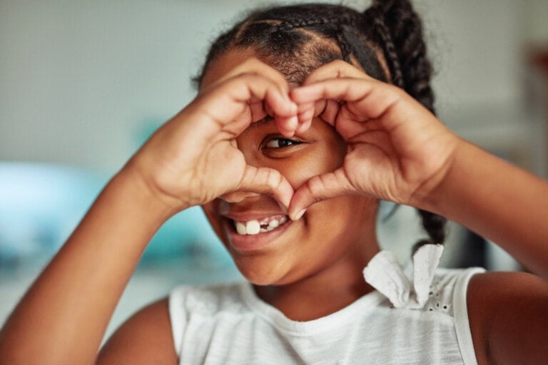 A loving child with braided hair is smiling and holding her hands in the shape of a heart over her face. She is wearing a white sleeveless top with a folded piece of paper peeking out from the strap. The background is blurred.