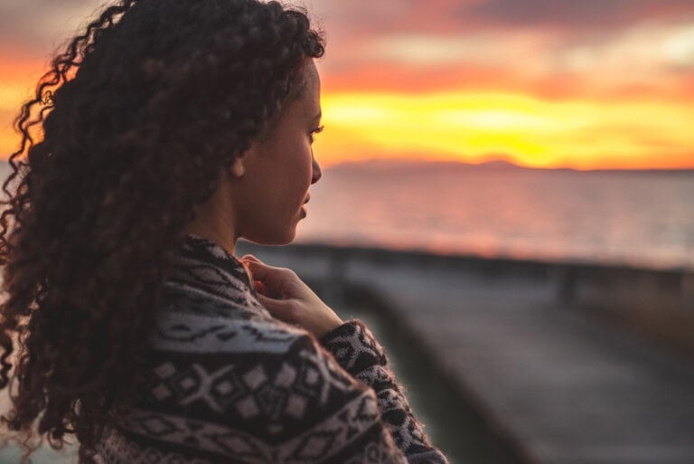 Young African American woman is standing on the promenade at the lake, looking thoughtfully towards the water and the setting sun. The girl in a vest is in a thoughtful mood.