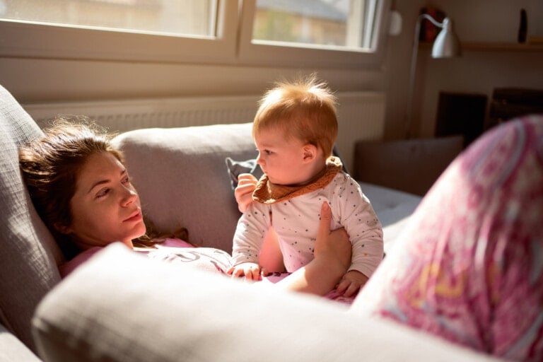 A woman is lying on a couch looking at a baby who is sitting on her lap. Sunlight is streaming through the window behind them, creating a cozy atmosphere. Despite the serene scene, scary thoughts new moms have hover in her mind as the baby holds a piece of cloth to their mouth while gazing back at her.
