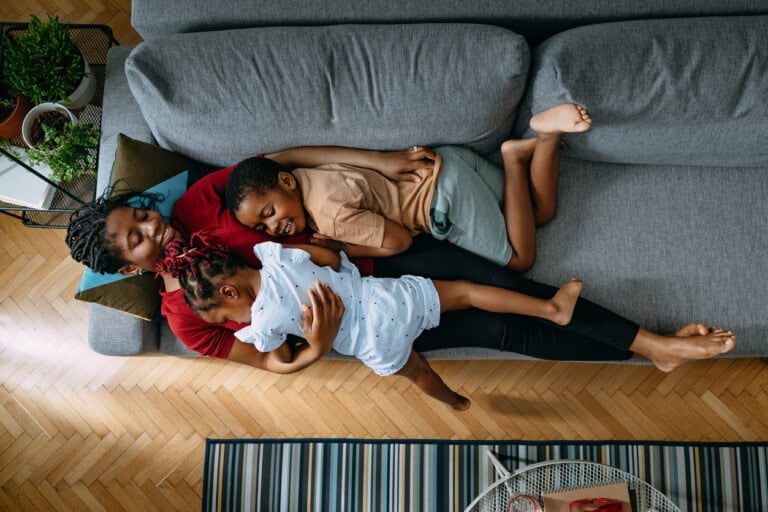 A scene of motherhood unfolds as a person and two children lie on a gray couch. The person holds one child close, embodying tenderness, while the other child rests peacefully beside them. The room, adorned with potted plants, has a warm wooden floor that enhances the cozy atmosphere.