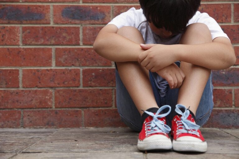 A young child is sitting on the ground with their head down and arms crossed over their knees. They are wearing a short-sleeved shirt, shorts, and red sneakers with blue laces. The background consists of a brick wall, symbolizing the importance of learning from mistakes and accepting failure.
