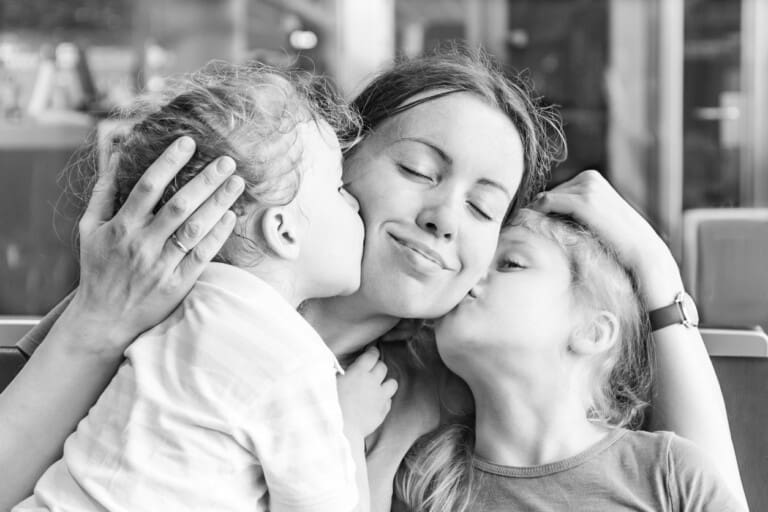 A black-and-white photo features a woman seated and smiling as two children, one on either side, kiss her cheeks. The woman gently holds each child with her hands. Despite any past birth trauma, all three appear happy and affectionate in this candid moment.