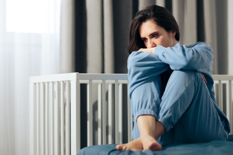 A person wearing blue pajamas sits on a bed with knees pulled up, their arms wrapped around their legs. They appear contemplative or worried, possibly having a miscarriage. Behind them, there's a white crib and grey curtains hanging in the background.
