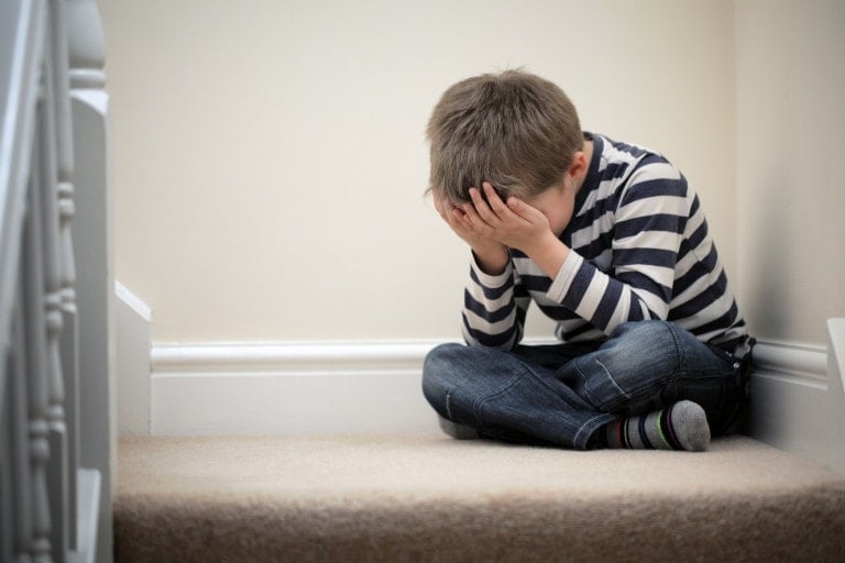 A young boy sits on a carpeted staircase with his face buried in his hands. Wearing a striped long-sleeve shirt, jeans, and mismatched socks, he appears distressed or upset. The staircase and walls are painted white, creating a serene backdrop as efforts to help kids manage stress become essential.