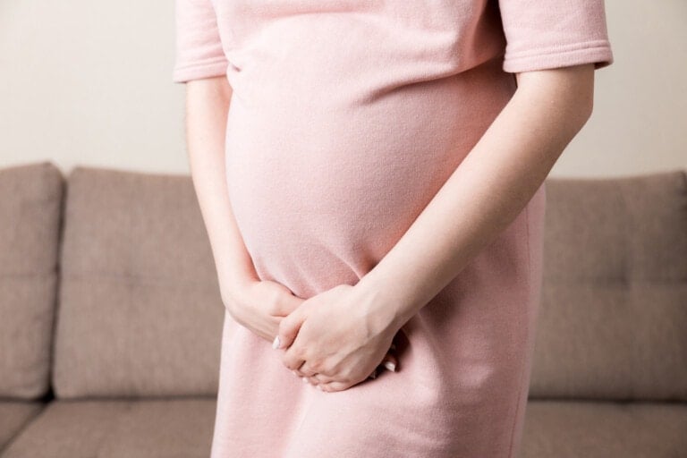 A person in a light pink dress holds their pregnant belly with both hands, standing in a room with a brown upholstered couch in the background. The upper part of their body and head are not visible, highlighting the importance of considering UTI while pregnant.