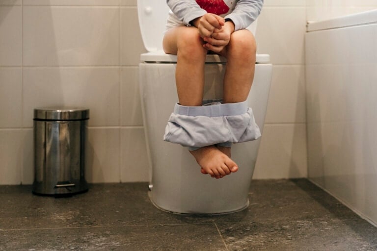 A child, dealing with childhood constipation, sits on a toilet with their pants around their ankles. They wear a long-sleeve top, and the bathroom has a trash bin to the left and white tiled walls. The image only captures the child from the chest down.