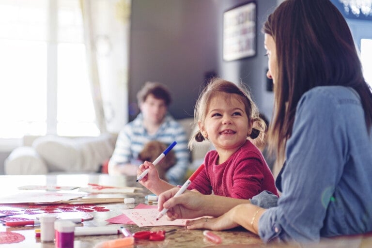 A young woman and a small child are sitting at a table drawing with markers, creating Valentine's Day crafts for kids. The child is smiling and looking at the woman. Art supplies and heart-shaped cutouts are scattered on the table. A man and a dog are seated on a couch in the background.