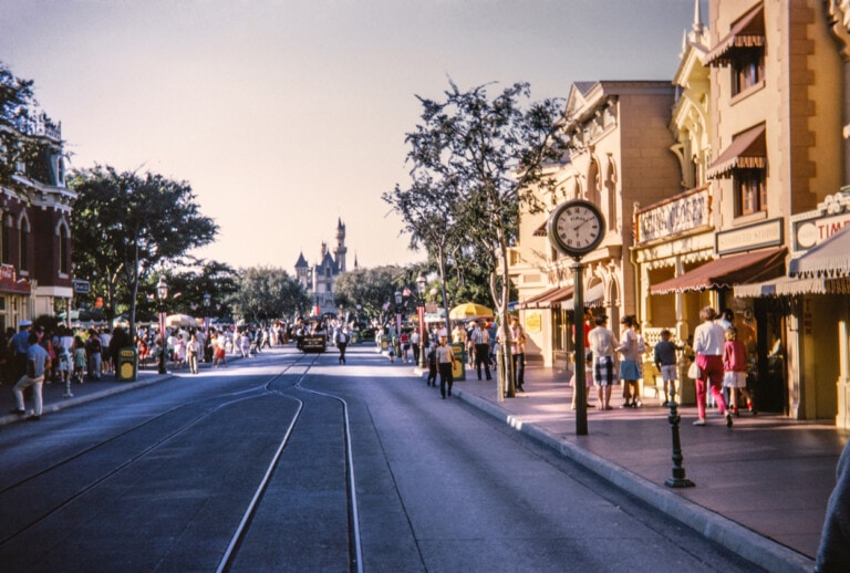 A street scene in the Magic Kingdom features a clock post and shops on either side, with people strolling around. In the background, a castle structure is visible. Tracks run down the middle of the street, leading to exciting Magic Kingdom rides. Various trees line the pathway.