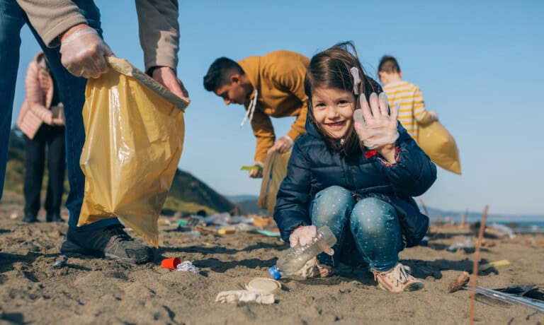 A young girl smiling and holding a plastic bottle while participating in a beach cleanup. She is wearing gloves and is surrounded by other volunteers picking up litter—one person holds a yellow bag. The sky is clear, and the beach is scattered with trash, showcasing random acts of kindness in action.