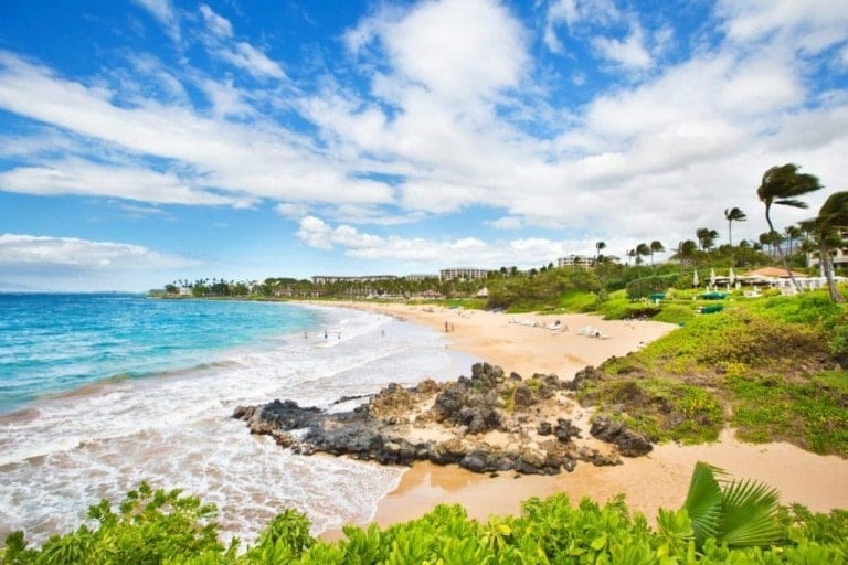 A scenic beach in Maui with golden sand and clear blue waters. People are seen enjoying the beach, with some swimming and others relaxing on the sand. Lush green vegetation borders the beach, and some buildings are visible in the background under a partly cloudy sky—perfect for a Maui babymoon.