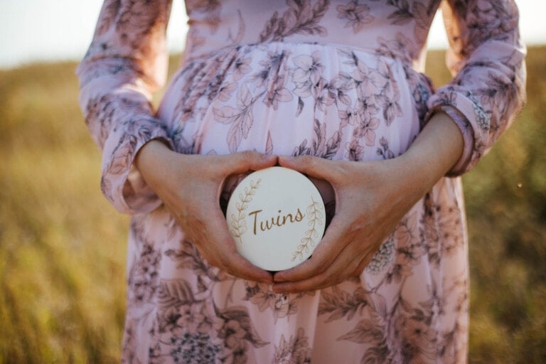 A person wearing a floral dress is outdoors, holding a round sign with the word "Twins" in gold lettering, positioned in front of their visibly pregnant belly. The background is a grassy field with warm sunlight, capturing the joyous moment after learning how to get pregnant with twins.