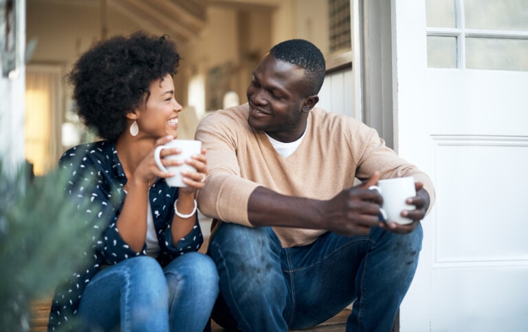 You and your partner are sitting on a porch, each holding a white mug. You're smiling at each other. The woman has curly hair and is wearing a blue polka-dotted shirt. The man has short hair and is wearing a beige sweater and jeans. A door is open behind you both.
