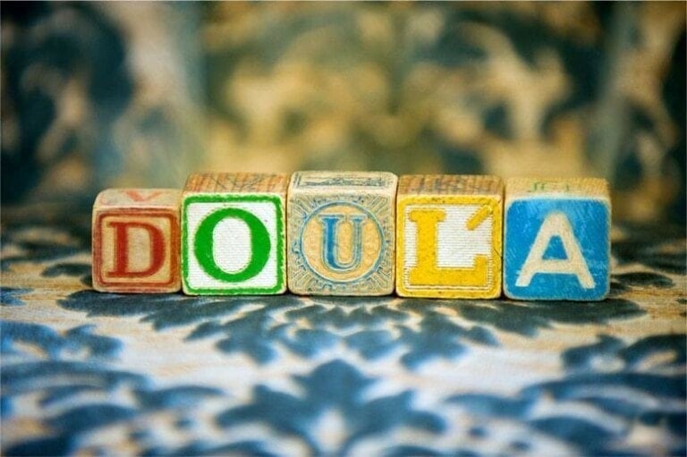 Five colorful letter blocks are arranged to spell DOULA on a patterned surface, prompting the question: what does doula mean?