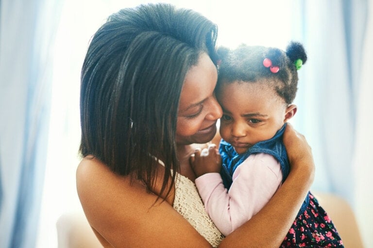 An adult holding a young child, who appears upset from toddler tantrums, in a comforting embrace. The adult has long hair and is wearing a light-colored sleeveless top. The child, with their hair in two pigtails featuring colorful accessories, is wearing a denim vest over a pink shirt.