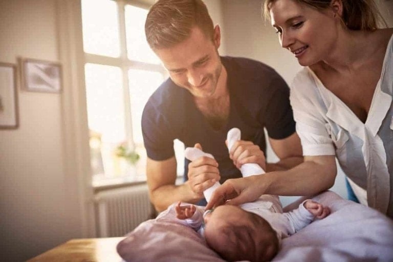 A man and a woman are smiling and tending to a baby on a changing table. The man is holding the baby's legs while the woman gently touches the baby's stomach. They are in a well-lit room with framed pictures on the wall, showcasing how dads can help a new mom with care routines.