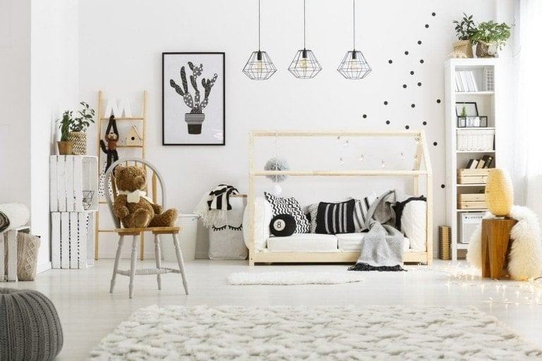 A well-lit, modern, gender-neutral nursery features a wooden bed with black and white bedding, a stuffed bear on a chair, geometric hanging lights, and decor including a cactus poster and polka dot wall decals. The room is decorated in neutral tones with cozy elements.