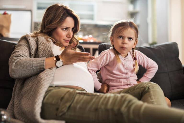 A pregnant woman sits on a couch next to a young girl. The woman, dressed in a gray cardigan and green pants, gestures with her hand while looking at the child. The young girl, wearing a pink cardigan and pigtails, has a concerned expression on her face as if theyre discussing how to handle back talk.