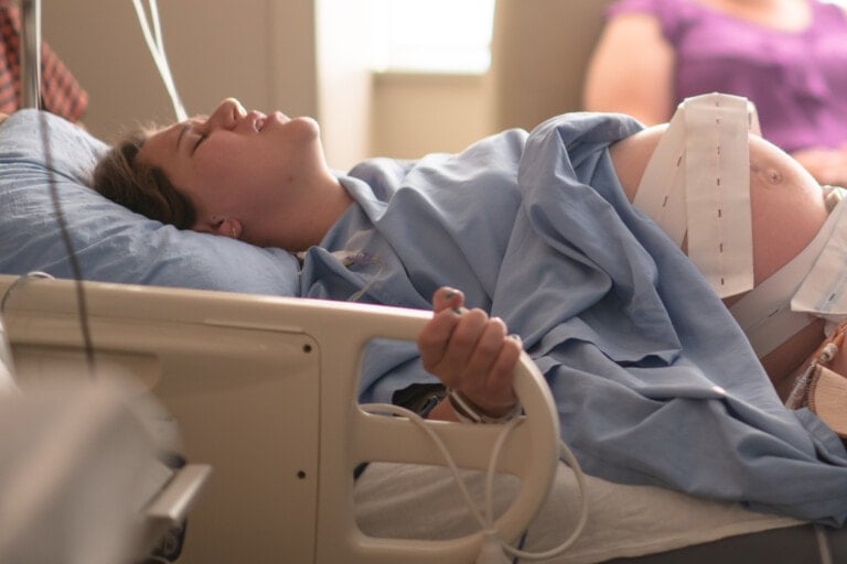 A woman in labor lies in a hospital bed wearing a blue gown, with monitoring equipment attached to her abdomen. Her face shows signs of effort as she lets out a deep groan. A person in a purple shirt is partially visible in the background, seated next to the bed.