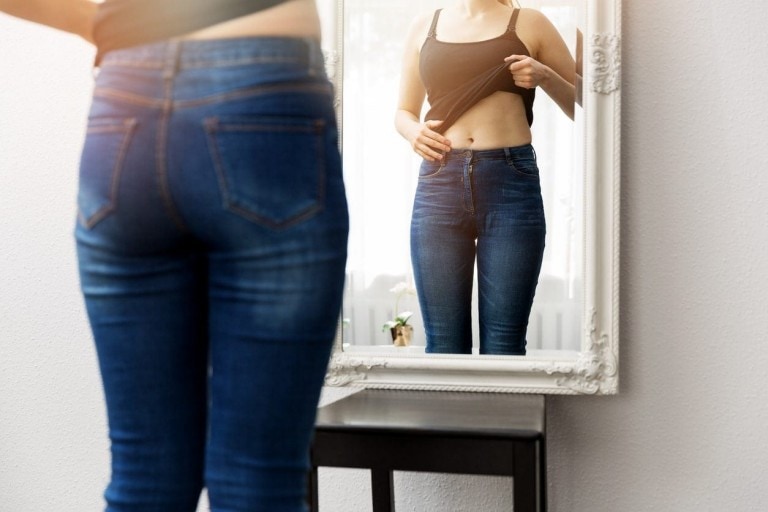 A person wearing a black top and blue jeans stands in front of a mirror, lifting their top slightly and examining their reflection. The mirror has a white frame, and a small plant is visible in the background. They seem to be observing weight loss during their menstrual cycle.