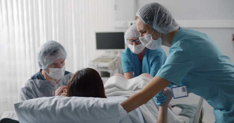 Three healthcare workers in scrubs, masks, and hair covers are attending to a patient lying in a hospital bed. One worker appears to be comforting the patient, perhaps managing an instance of shoulder dystocia, while the other two look on. The room contains medical equipment and a window with blinds.