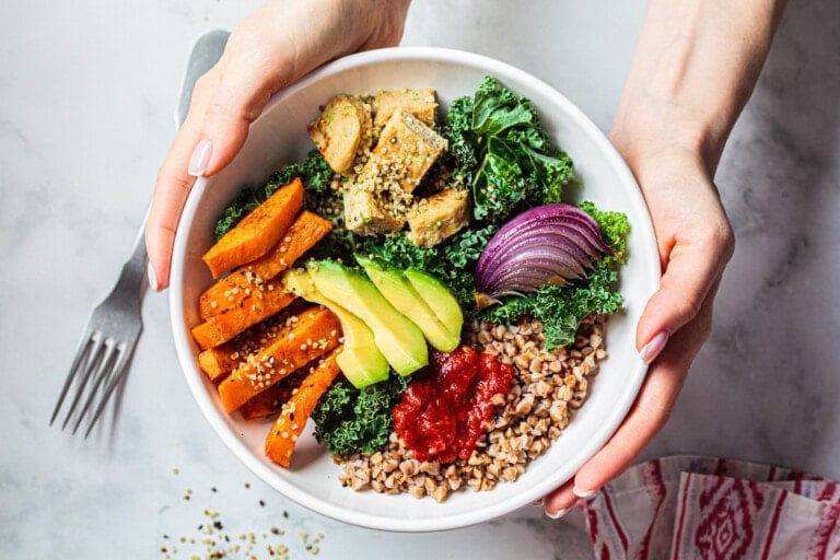A person is holding a white bowl containing a healthy salad, perfect for maintaining a vegan diet during pregnancy. The salad includes kale, sliced avocado, sweet potato wedges, red onion, buckwheat, and a dollop of red sauce. A fork and a patterned napkin are partially visible in the background.