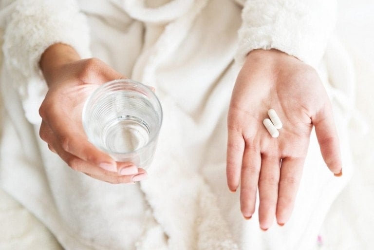 A person wearing a white robe holds a glass of water in their right hand and two white postpartum vitamins in their left hand. The image is a close-up, focusing on the hands and the objects they are holding.