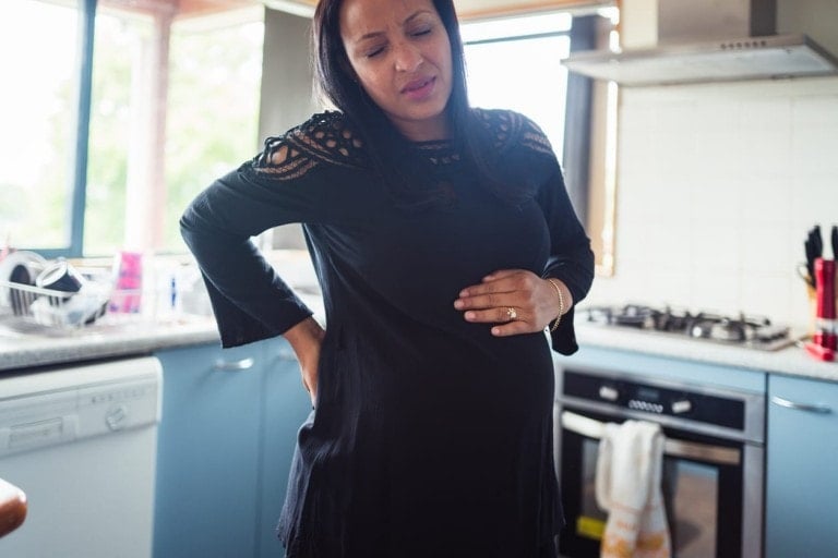 A pregnant woman is standing in a kitchen, wearing a black dress with lace details. With one hand on her lower back and the other on her belly, she appears to be experiencing pelvic pain during pregnancy. Kitchen appliances, including a dishwasher and stove, are visible in the background.