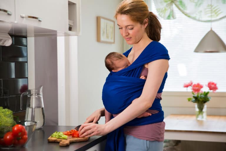 A woman in a kitchen is preparing food while carrying a baby in a blue wrap. She is slicing a red bell pepper on a cutting board. A vase of pink flowers is on the table, and various vegetables are on the counter.