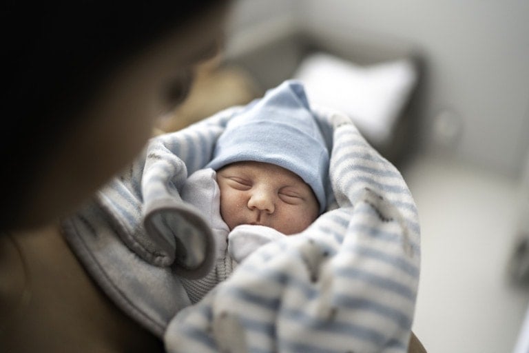 A newborn baby, who just underwent a circumcision, is wrapped in a blue and white striped blanket, wearing a blue hat. The baby is sleeping peacefully, held by an adult whose face is partially visible. The background is blurred.