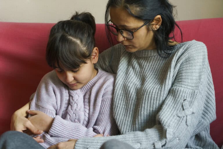 A woman wearing glasses and a gray sweater sits closely with her daughter, a young girl in a pink sweater on a red couch. The woman is hugging the girl and both appear to be in a solemn moment.