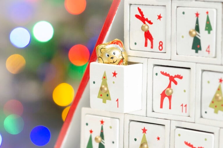 A close-up of a Christmas advent calendar with small white drawers. One drawer is open, revealing a gold foil-wrapped chocolate bear. The drawers are decorated with Christmas-themed images like trees and reindeer. Colorful lights are blurred in the background.