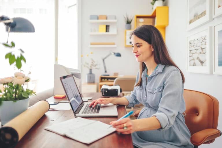 A woman on maternity leave sits at a desk in her home office, working on a laptop and writing in a notebook. The workspace features a plant, rolled paper, and office supplies, with shelves and framed pictures decorating the background.