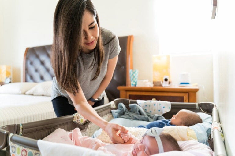 A woman stands near a crib, gently touching one of two infants lying inside. The crib is positioned next to a bed and a wooden nightstand with a lamp and other small items on it. The room is well-lit with natural light coming from a window, showcasing some of the best baby products for twins.