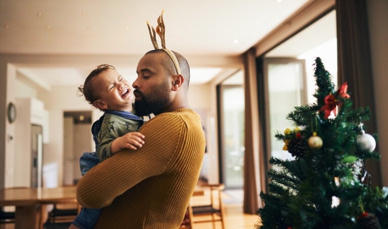 A man wearing a mustard yellow sweater and reindeer antlers kisses a smiling toddler, with no tantrums during the holidays in sight. They are standing next to a decorated Christmas tree in a bright, modern living room. The toddler is wearing denim overalls.