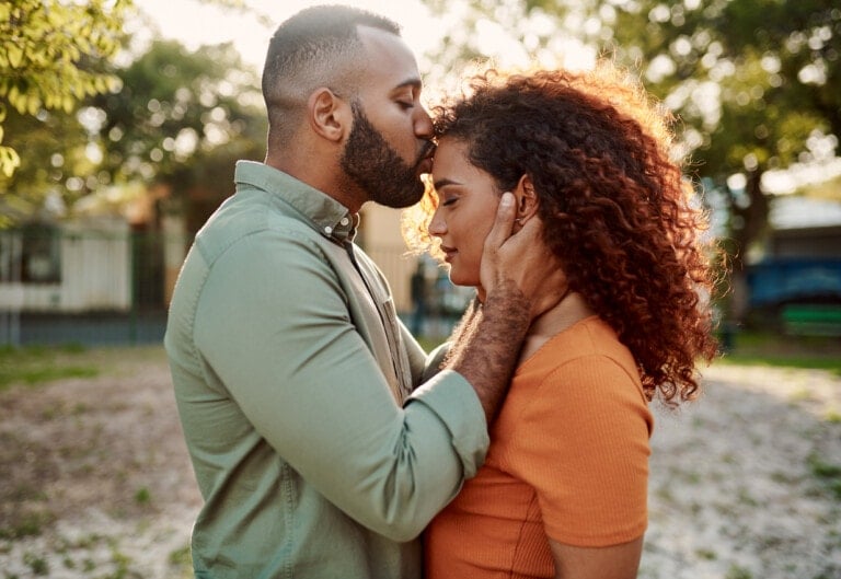 A man with short hair and a beard gently kisses a woman with curly hair on the forehead during their date night. He cradles her head with both hands as she closes her eyes, appearing calm. They are outdoors with trees and a blurred background in a sunlit area.