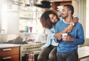 A woman sits on a kitchen counter, smiling and hugging a man holding a coffee mug. They are both casually dressed and appear happy in their bright, modern kitchen. Various kitchen items and appliances are visible in the background as they enjoy a moment of togetherness, reflecting their split responsibilities.