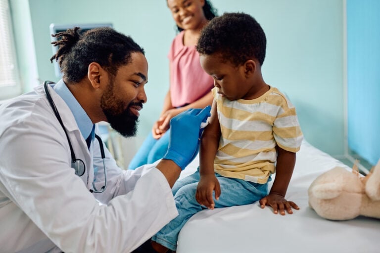 A healthcare professional with a beard and gloves is administering a shot to a young child on a medical examination table. The child's clothing consists of a yellow and white striped shirt and jeans. An adult is sitting nearby, smiling and observing the process of how to get over fear of needles.