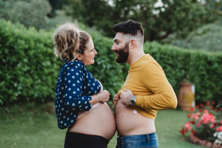 Two people stand outdoors, each pulling up their shirts to expose their bellies. The person on the left has a visibly pregnant belly and is smiling at the person on the right, who is not pregnant but mimics the pose. It's likely part of a playful dadchelor party in a garden with greenery in the background.