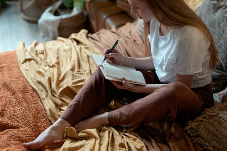 A person with long hair is sitting on a bed with beige and orange blankets, immersed in writing about the benefits of journaling. The individual is wearing a white shirt and brown pants. The setting appears to be a cozy, rustic room with woven baskets in the background.