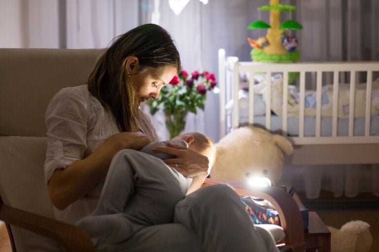 A woman sits in a chair holding a baby in a dimly lit room, perhaps contemplating gentle night weaning. Behind them, there is a crib with stuffed toys and a large teddy bear on the floor. A bouquet of flowers is seen near the window.